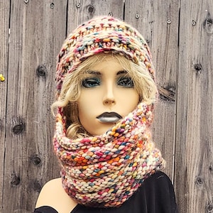 Puede incluir: Un conjunto de gorro y cuello tejidos a mano, de colores. El gorro y el cuello están hechos de hilo grueso y texturizado en una mezcla de crema, rosa, morado, azul y amarillo. El cuello está alrededor del cuello de un maniquí.