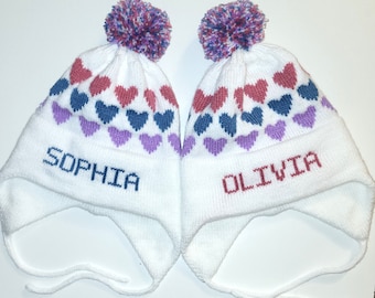 Puede incluir: Dos gorros de invierno de punto blanco con pompones, con un patrón de corazones rosas, morados y azules. Los gorros tienen los nombres "Sophia" y "Olivia" bordados.