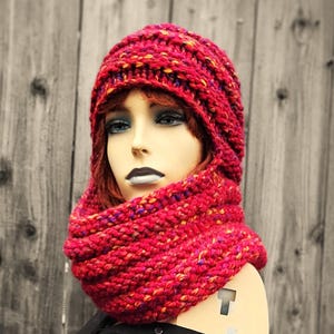 May include: A vibrant red knit hat and cowl set. The hat is a close-fitting beanie style, and the cowl is a wide loop scarf. Both pieces are made of a chunky knit with flecks of yellow, orange, and purple. The set is displayed on a mannequin head.