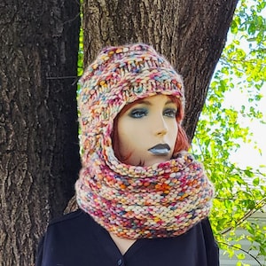 May include: A chunky knit balaclava featuring a multicolored yarn blend of pink, orange, yellow, blue, and cream tones. The headpiece has a thick ribbed texture and covers the head and neck, framing the face with a wide, circular opening.