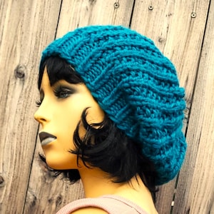 Könnte beinhalten: Eine türkisfarbene Strickmütze mit einem lockeren Sitz. Die Mütze ist aus weichem, dickem Garn gefertigt und hat ein schlichtes, klassisches Design.