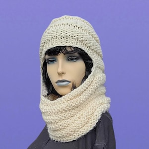 May include: A cream-colored knitted hood and cowl set. The hood covers the head and frames the face, while the cowl wraps around the neck. The knitwear has a textured appearance, suggesting a handmade or artisanal quality. The set is displayed on a mannequin.