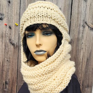 May include: Cream-colored knitted hat and cowl set. The hat has a rounded crown and a face-framing design. The cowl is a wide, knitted tube. The set is displayed on a mannequin with black hair and blue eye makeup. The background is a wooden wall.