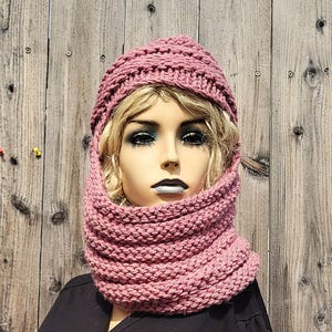 May include: A dusty rose-colored knitted hat and cowl set. The hat is a beanie style, and the cowl is a long, looped scarf. The set is made of a thick, textured yarn, perfect for cold weather. The set is displayed on a mannequin.