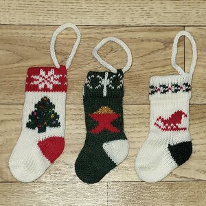 May include: Three miniature knitted Christmas stockings with white hanging loops. One stocking features a red and white snowflake design, another has a green Christmas tree, and the third has a red star. The stockings are white, red, and green.