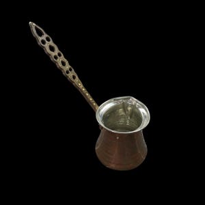 May include: A copper coffee pot with a long, ornate handle. The handle has a decorative pattern with diamond-shaped cutouts. The pot has a silver-colored interior and a spout for pouring. The pot is designed for making coffee.
