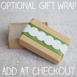 May include: A brown cardboard gift box with a green ribbon and white scalloped trim. The box is open and the inside is lined with white tissue paper. The text "OPTIONAL GIFT WRAP" is at the top of the image and "ADD AT CHECKOUT" is at the bottom.