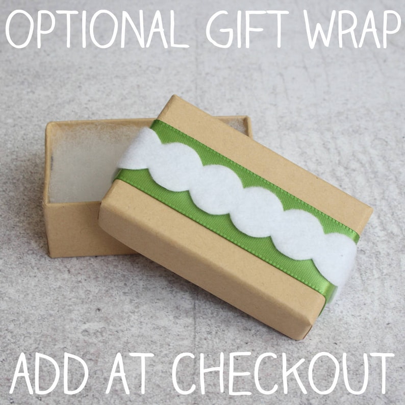 May include: A brown cardboard gift box with a green ribbon and white scalloped trim. The box is open and the inside is lined with white tissue paper. The text "OPTIONAL GIFT WRAP" is at the top of the image and "ADD AT CHECKOUT" is at the bottom.