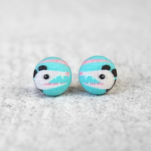 May include: A pair of small, round, turquoise fabric earrings with a panda bear design. The panda bears have black eyes and pink cheeks.