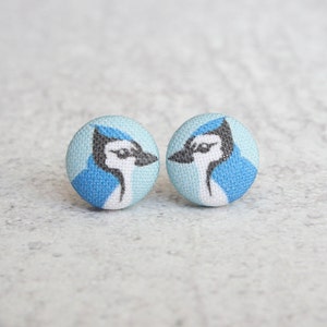 May include: A pair of blue jay stud earrings. The earrings are made of fabric with a blue jay design on a light blue background.