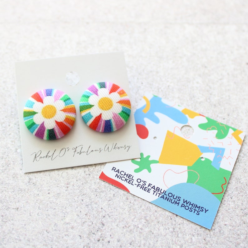 May include: A pair of colorful rainbow and white daisy fabric button earrings with nickel-free titanium posts. The earrings are displayed on a white card with the text "Rachel O's Fabulous Whimsy" and a separate card with the text "Rachel O's Fabulous Whimsy Nickel-Free Titanium Posts".