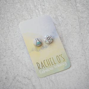 May include: A pair of science-themed stud earrings. One earring features a beaker with blue liquid, and the other earring features an atom with a white center and black lines. The earrings are on a white card with the text "Rachel O's fabulous whimsy".