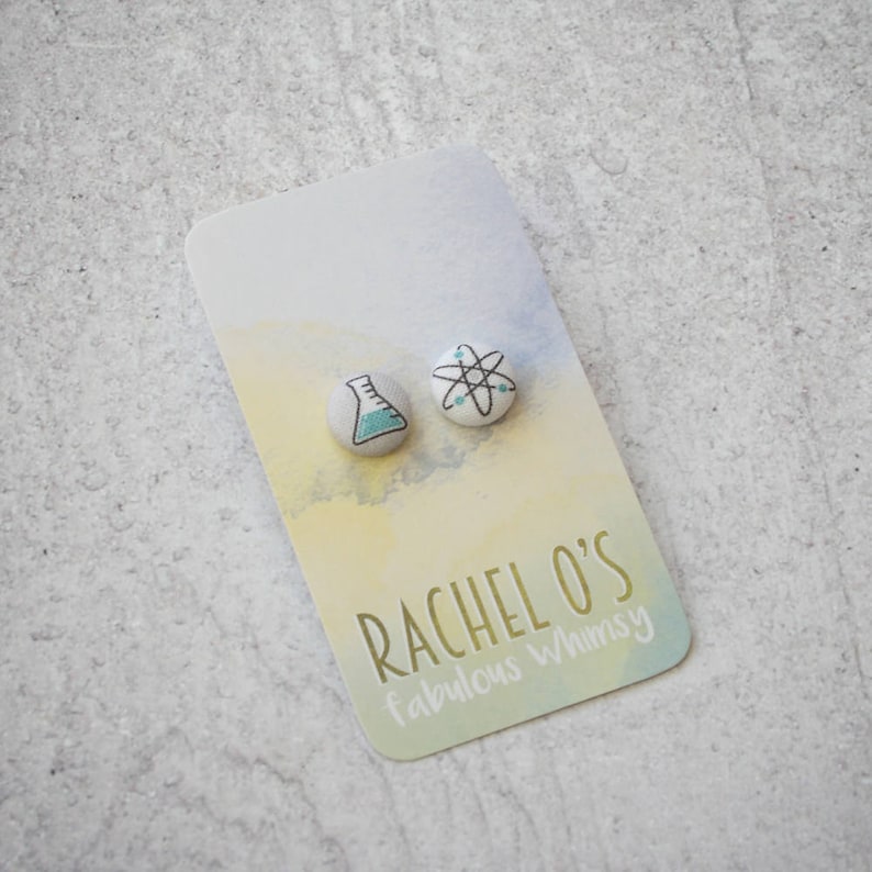 May include: A pair of science-themed stud earrings. One earring features a beaker with blue liquid, and the other earring features an atom with a white center and black lines. The earrings are on a white card with the text "Rachel O's fabulous whimsy".