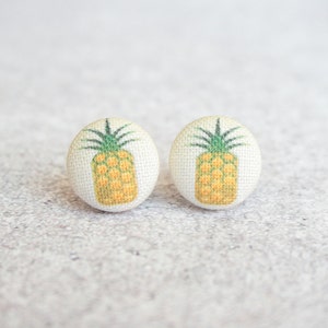 May include: A pair of white fabric button earrings with a pineapple design. The pineapples are yellow with green tops.