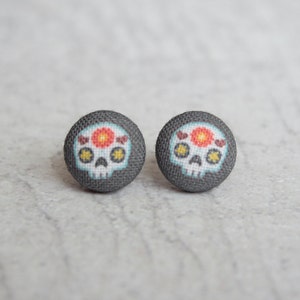 May include: A pair of gray fabric stud earrings with a white sugar skull design. The skull has a red flower and yellow stars.