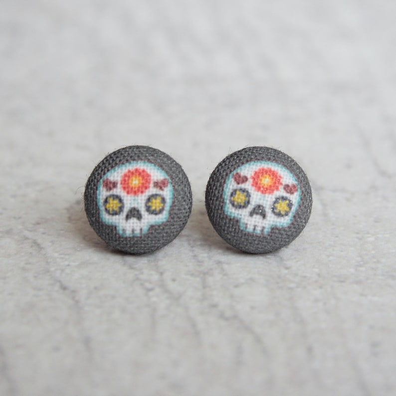 May include: A pair of gray fabric stud earrings with a white sugar skull design. The skull has a red flower and yellow stars.