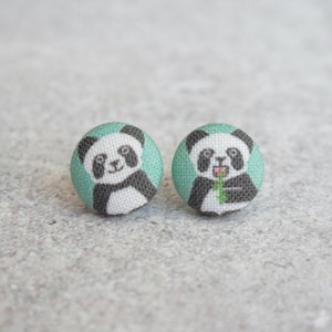 May include: A pair of panda bear stud earrings. The earrings are made of fabric and have a teal background. One panda is holding a piece of bamboo.