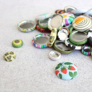 May include: A collection of colorful fabric covered buttons with different patterns, including a sunflower, leopard print, and strawberries. The buttons are arranged on a light gray surface.