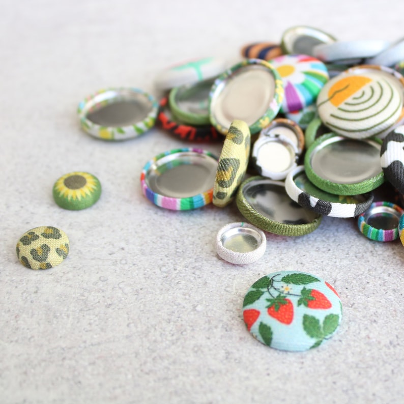 May include: A collection of colorful fabric covered buttons with different patterns, including a sunflower, leopard print, and strawberries. The buttons are arranged on a light gray surface.