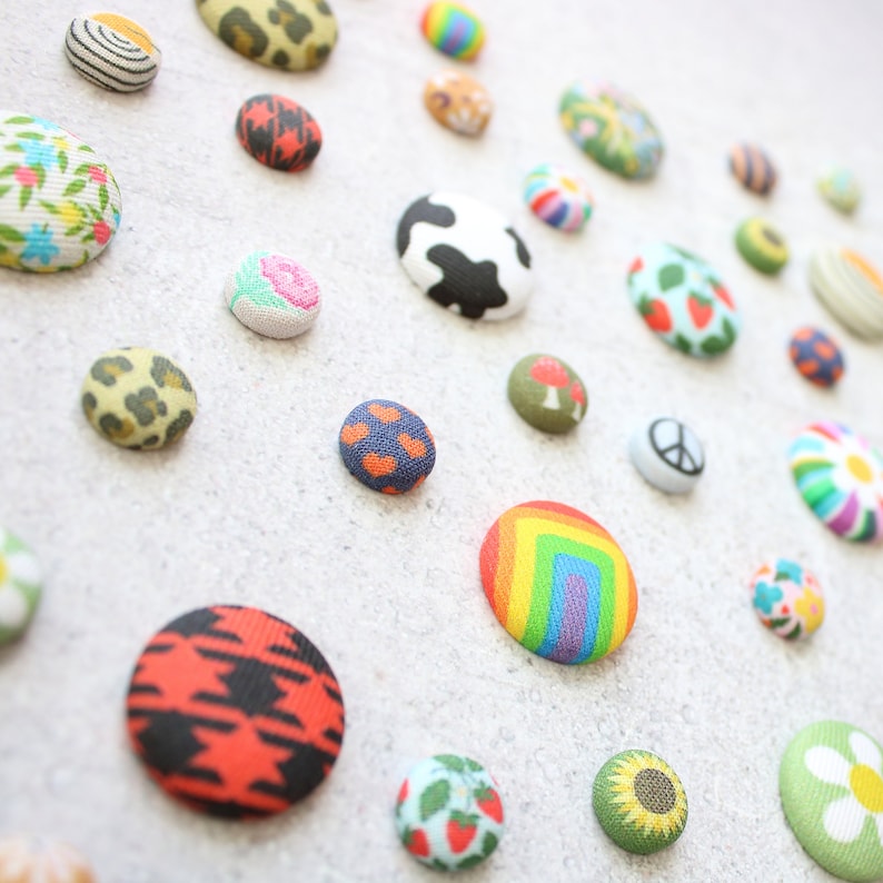 May include: A collection of colorful fabric buttons with various patterns, including a cow, a peace sign, a rainbow, a sunflower, and a mushroom.