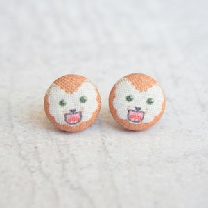 May include: A pair of small, round, fabric earrings with a cartoon animal face design. The earrings are peach colored with a white face and green eyes. The animal has a pink mouth and a small black nose.