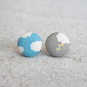 May include: Two round fabric buttons. One button is light blue with white cloud shapes. The other is gray with a white cloud and yellow lightning bolt design. These buttons are perfect for sewing and craft projects.