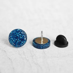 May include: Three-piece set of blue glitter push pins. One pin is fully assembled, showing a round, glitter-covered head and a silver pin. The second pin shows the pin's components, and the third is the black backing.