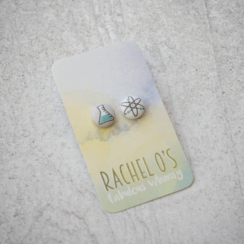 May include: A pair of science-themed stud earrings. One earring features a beaker with blue liquid, and the other earring features an atom with a white center and black lines. The earrings are on a white card with the text "Rachel O's fabulous whimsy".