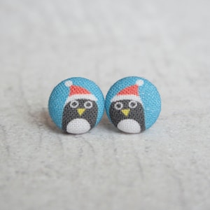 May include: A pair of blue fabric earrings with a penguin wearing a red Santa hat design.