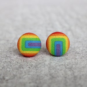 May include: A pair of rainbow-colored fabric button earrings with a concentric square design. The earrings are a vibrant mix of red, orange, yellow, green, blue, indigo, and violet.