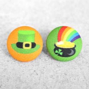 May include: Two round fabric buttons with a St. Patrick's Day theme. One button features a green leprechaun hat with a gold buckle on an orange background. The other button features a green pot of gold with a rainbow on a green background.