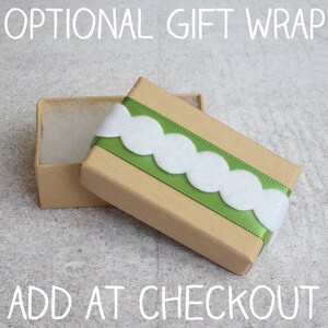 May include: A brown cardboard gift box with a green ribbon and white scalloped trim. The box is open and the inside is lined with white tissue paper. The text "OPTIONAL GIFT WRAP" is at the top of the image and "ADD AT CHECKOUT" is at the bottom.