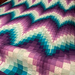 Può includere: Una colorata trapunta con un design geometrico nelle tonalità del viola, del verde acqua e del bianco. La trapunta presenta un motivo a zig-zag ed è realizzata con quadrati. La trapunta è su un letto e ha una texture trapuntata.