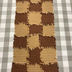 Könnte beinhalten: Ein rechteckiger Tischläufer mit Patchwork-Design. Abwechselnd Quadrate aus braunem Karostoff und beige gestreiftem Stoff sind zusammengenäht. Die Kanten sind ausgefranst, was einen rustikalen, hausgemachten Eindruck erweckt. Der Läufer ist wahrscheinlich aus Baumwolle.