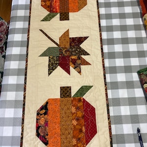 Könnte beinhalten: Ein beiger Tischläufer mit Patchwork-Muster, das zwei orangefarbene Kürbisse und ein rot-grün-braunes Blatt zeigt. Der Läufer hat einen braunen Rand.