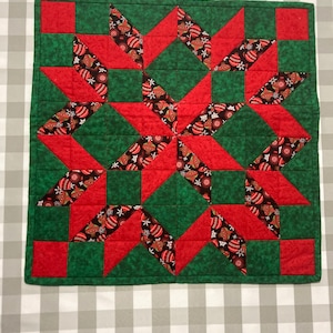 May include: A red and green quilted Christmas table runner with a starburst pattern. The fabric features a repeating pattern of ornaments and snowflakes.