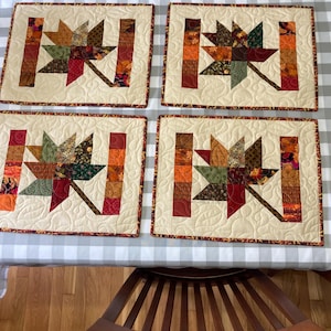 May include: Four quilted placemats with a maple leaf design in shades of brown, orange, red, and green. The placemats are on a white and gray checkered tablecloth.