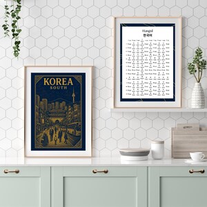 May include: Two framed prints on a white tiled wall. The print on the left is a blue and gold illustration of a city street scene with the text "KOREA SOUTH". The print on the right is a black and white chart with the text "Hangul 한글" at the top.