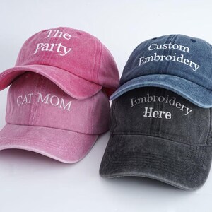 Pode incluir: Três bonés de beisebol em tons de rosa, azul e cinza. Os bonés rosa têm as frases "The Party" e "Cat Mom" bordadas em branco. Os bonés azuis e cinzentos têm "Custom Embroidery" e "Embroidery Here" bordados em branco.