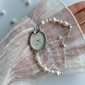 Op de afbeelding: Een zilverkleurig quartz horloge met een ovale wijzerplaat en een parelarmband. De wijzerplaat is wit met kleine kristallen en zilveren wijzers. De armband is gemaakt van onregelmatige parels en zilveren kralen.