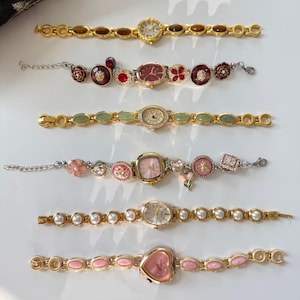 May include: Six decorative women's watches with various designs. The watches have gold-toned metal bands and feature different face shapes, including round, oval, and heart-shaped. Some watches have gemstone or pearl accents, and others have floral or decorative elements.