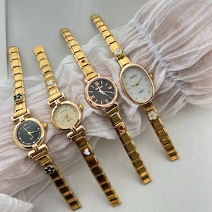 Op de afbeelding: Vier goudkleurige polshorloges met verschillende wijzerplaatontwerpen. De horloges hebben een goudkleurige metalen band met kleine decoratieve bedels. De horloges hebben ronde wijzerplaten met zwarte, witte en cr&egrave;mekleurige wijzerplaten. De horloges hebben Romeinse cijfermarkeringen en zwarte wijzers.