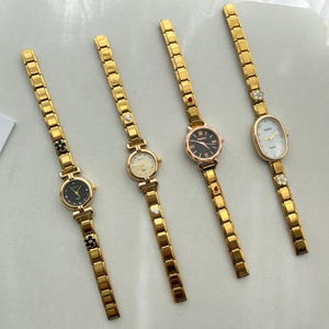 Op de afbeelding: Vier gouden pols horloges met verschillende wijzerplaatontwerpen. Elk horloge heeft een gouden metalen band met decoratieve accenten.