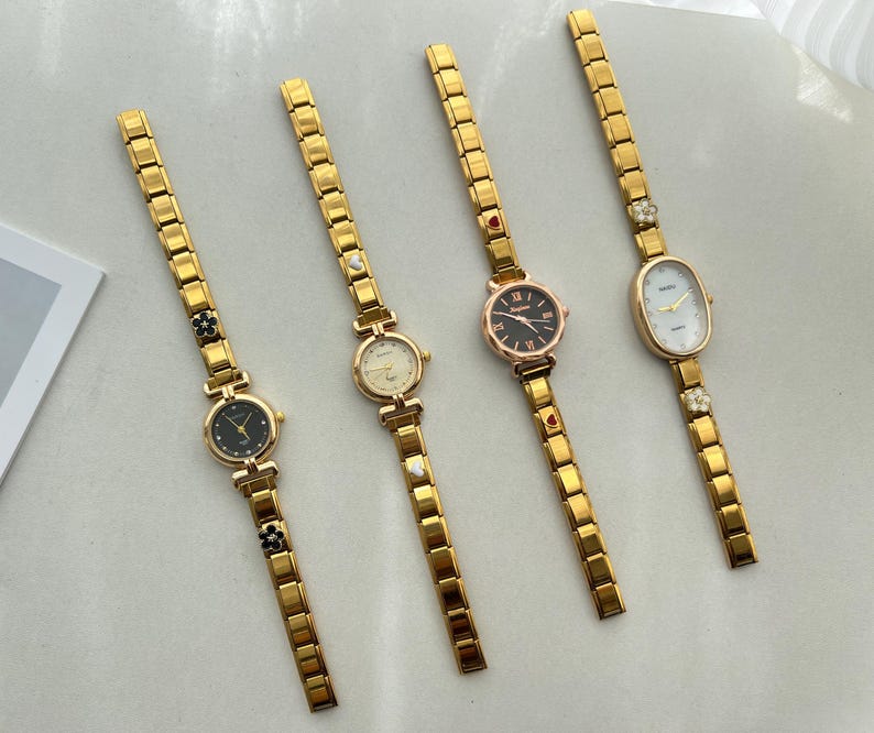 Op de afbeelding: Vier gouden pols horloges met verschillende wijzerplaatontwerpen. Elk horloge heeft een gouden metalen band met decoratieve accenten.