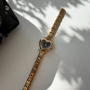 May include: A gold-toned wristwatch with a heart-shaped face and a black dial. The watch band is also gold and features a repeating star pattern. The watch is lying on a white surface next to a black fabric.