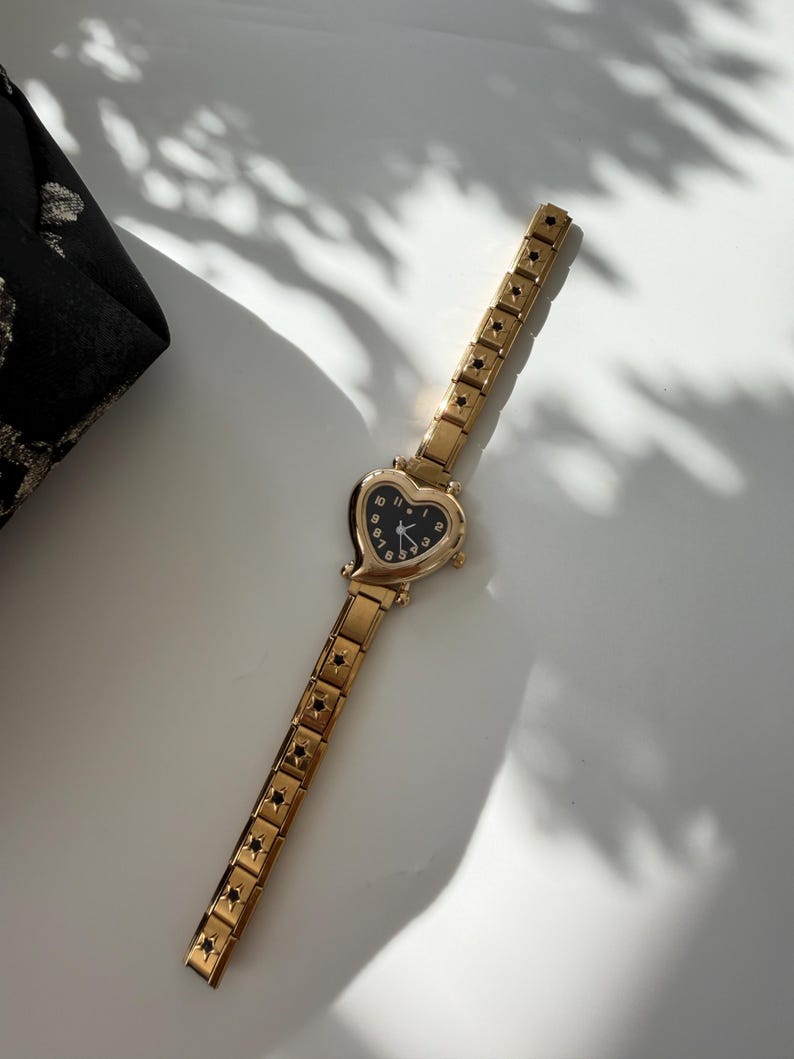 May include: A gold-toned wristwatch with a heart-shaped face and a black dial. The watch band is also gold and features a repeating star pattern. The watch is lying on a white surface next to a black fabric.