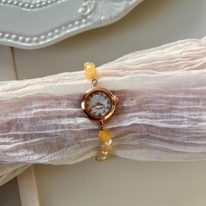 Pu&ograve; includere: Un orologio da polso in oro rosa con quadrante bianco e lancette dorate. L'orologio &egrave; attaccato a un braccialetto fatto di perline gialle. L'orologio e il braccialetto sono appoggiati su un tessuto rosa chiaro.