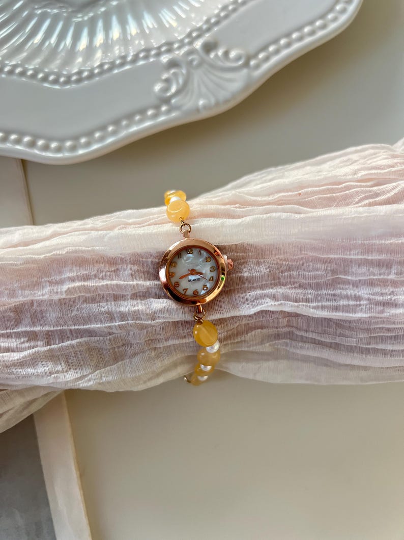 Pu&ograve; includere: Un orologio da polso in oro rosa con quadrante bianco e lancette dorate. L'orologio &egrave; attaccato a un braccialetto fatto di perline gialle. L'orologio e il braccialetto sono appoggiati su un tessuto rosa chiaro.
