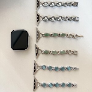 May include: Silver Apple Watch with a black screen, next to five watch bands. The bands feature silver links with oval-shaped embellishments in white, green, and blue. The bands are designed to be interchangeable.