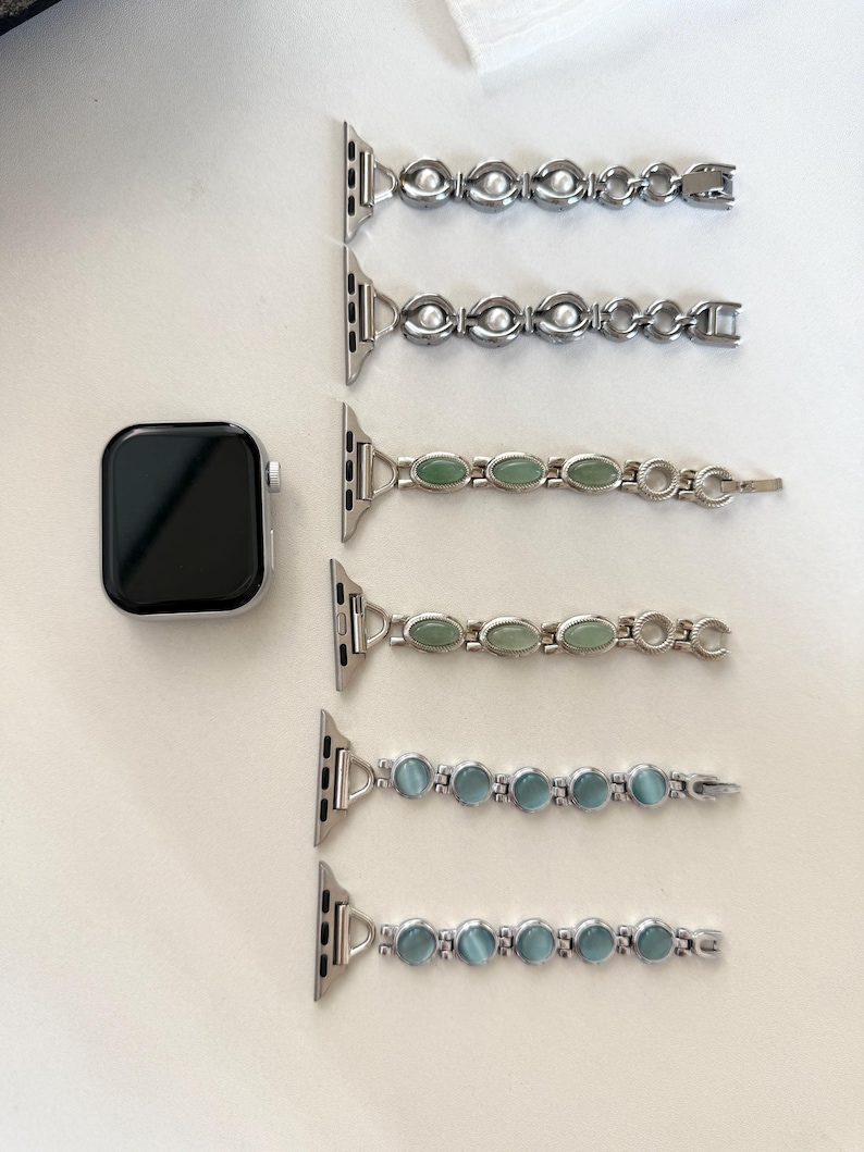 May include: Silver Apple Watch with a black screen, next to five watch bands. The bands feature silver links with oval-shaped embellishments in white, green, and blue. The bands are designed to be interchangeable.